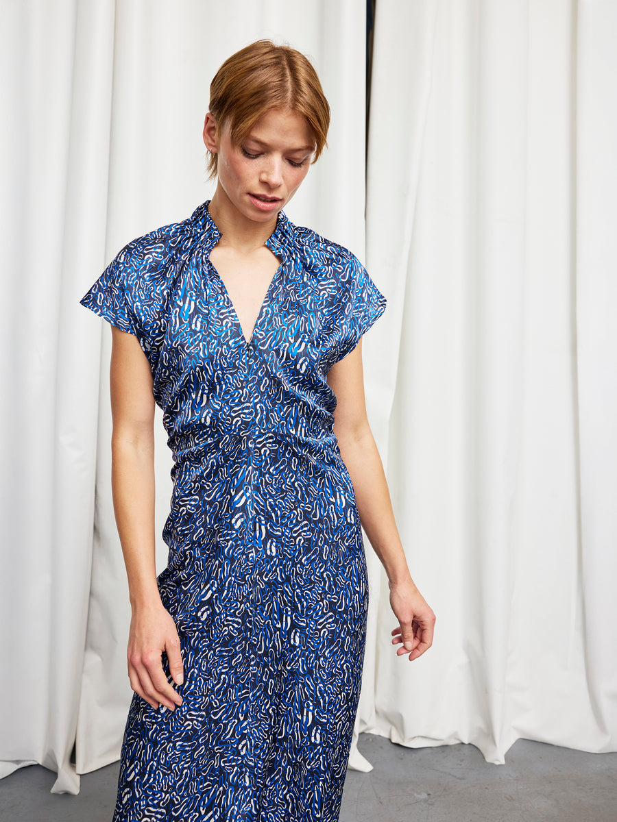 A person with short light brown hair stands indoors before white curtains, looking down with one hand at their side while wearing the Long Ruched Stella Dress by Zero + Maria Cornejo.