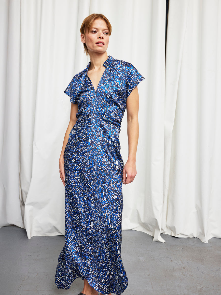 A woman with light brown hair pulled back stands before white curtains, wearing the Zero + Maria Cornejo Long Ruched Stella Dress—a blue, silk charmeuse full-length dress with short sleeves and a v-neck—looking relaxed to the side.