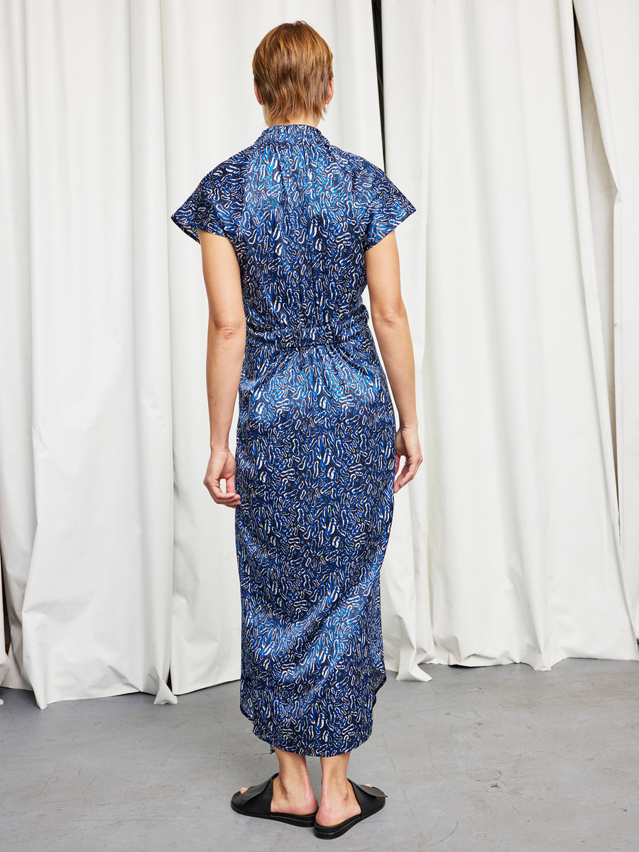 A person stands facing away, wearing the Zero + Maria Cornejo Long Ruched Stella Dress and black sandals, in front of white curtains on a gray floor.