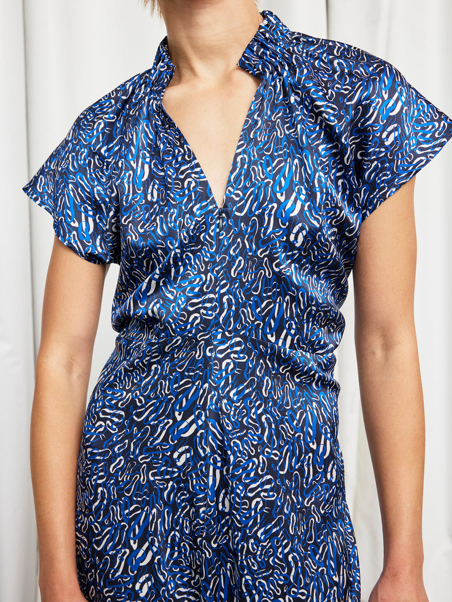 A person wearing the Zero + Maria Cornejo Long Ruched Stella Dress, a blue patterned silk charmeuse piece with short sleeves and a deep V-neckline, stands in front of a white curtain. Their head is not fully visible in the image.