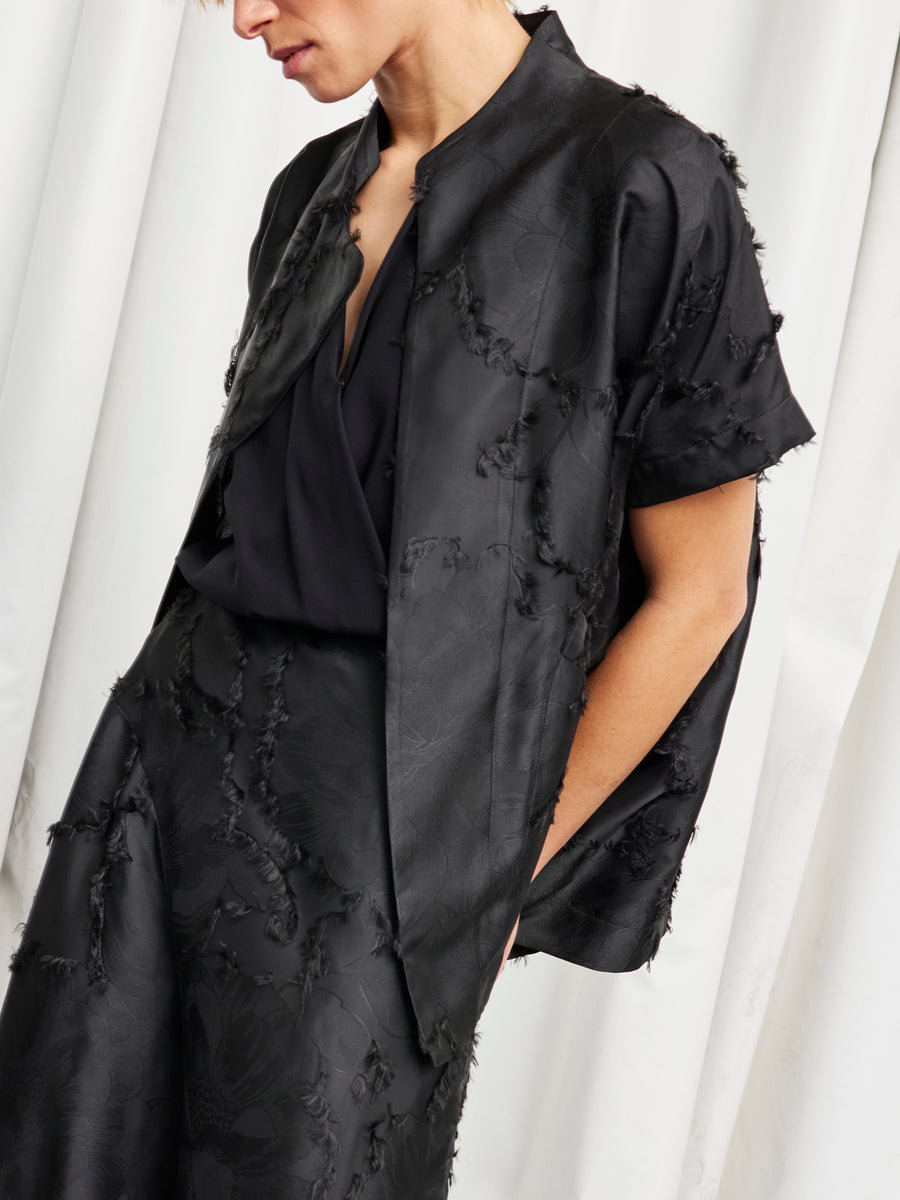 A person wears the Zero + Maria Cornejo Ari Shrug, a black floral fil coupe short-sleeve jacket with frayed edges, and a matching skirt, standing before a white curtain. Their face is partially out of frame. Both pieces are made in New York.