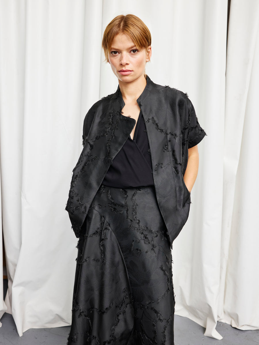 A woman with straight, light brown hair stands before white curtains in the black Ari Shrug by Zero + Maria Cornejo, hands in her pockets and looking directly at the camera.