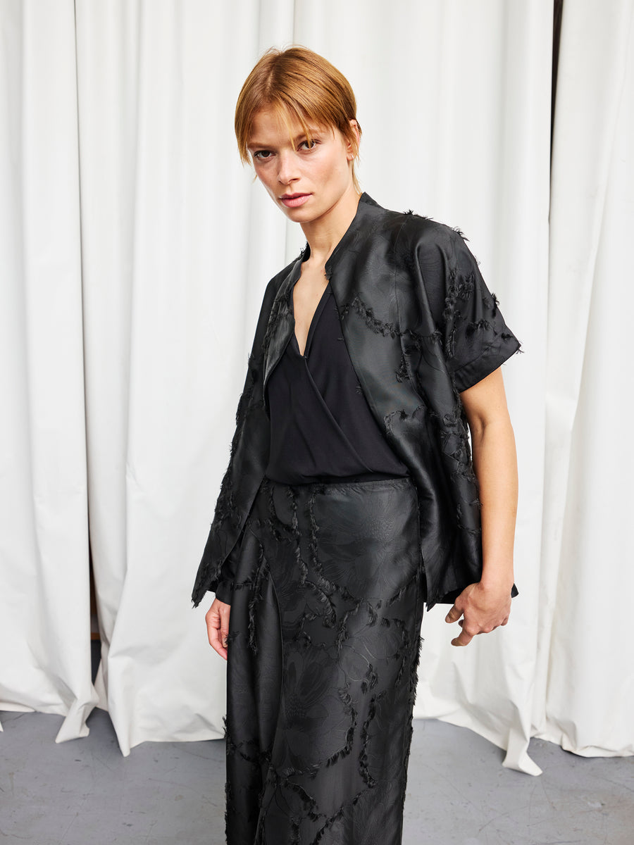 A person with short light brown hair wears the Ari Shrug by Zero + Maria Cornejo over a black top and textured black skirt, standing before white curtains and facing the camera.