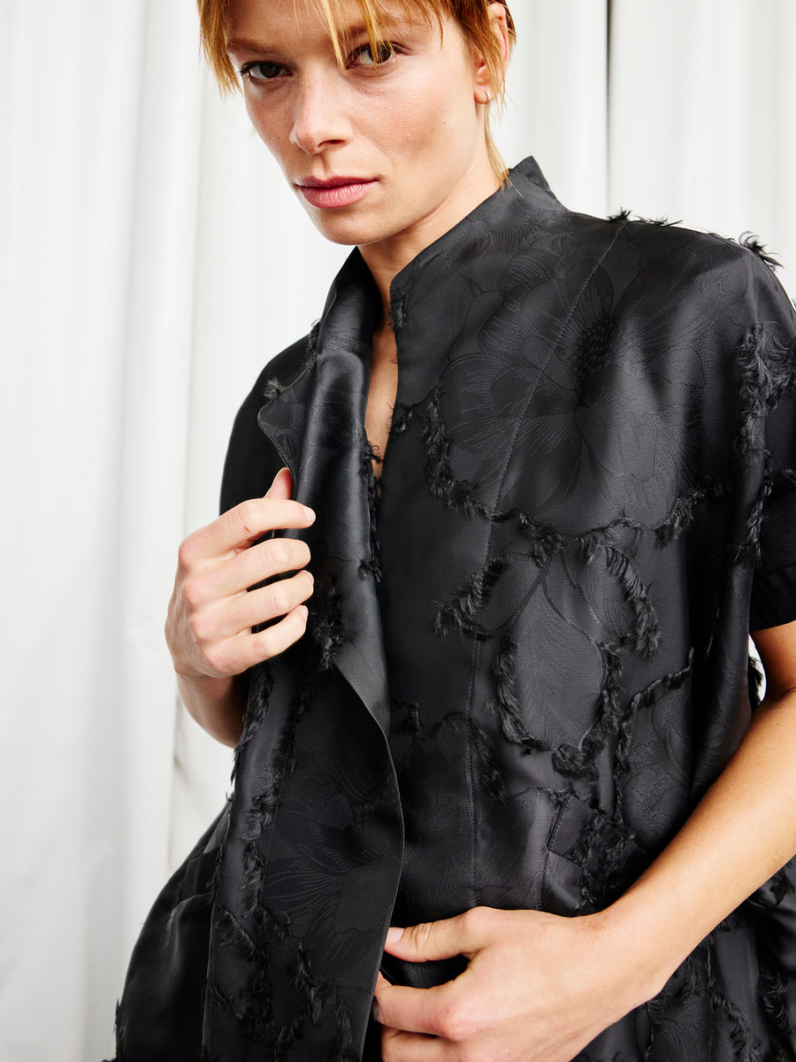 A person with short blond hair wears the Ari Shrug by Zero + Maria Cornejo, standing before white curtains and gazing intently at the camera while adjusting the collar with one hand.