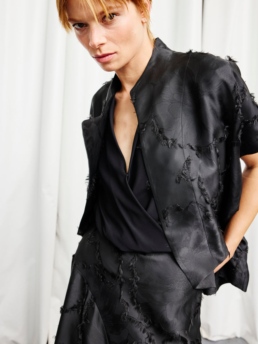 A person with short blonde hair wears the Ari Shrug by Zero + Maria Cornejo—a black textured jacket with subtle floral fil coupe patterns—paired with a matching skirt, standing before white curtains and gazing seriously at the camera.