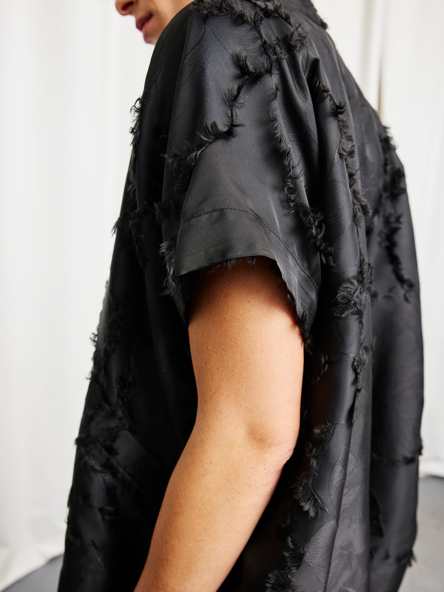 A person wears the Ari Shrug by Zero + Maria Cornejo—a short-sleeved, textured black piece with frayed details—against a white background, shown from shoulders to mid-arm with their face partially out of frame.