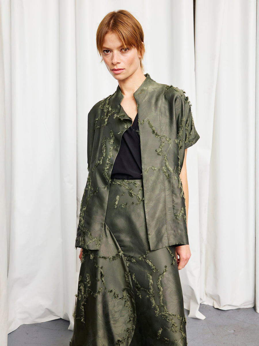 A person with light brown hair, wearing the Zero + Maria Cornejo Ari Shrug and a matching olive-green skirt over a black top, stands in front of white curtains.