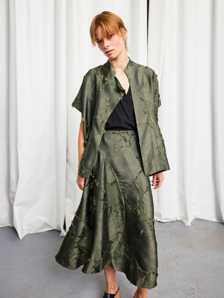 A person stands before white curtains, wearing the sustainable Ari Shrug by Zero + Maria Cornejo over a black top, paired with a textured olive green skirt and matching short-sleeve jacket. Their light brown hair is styled loosely and casually.