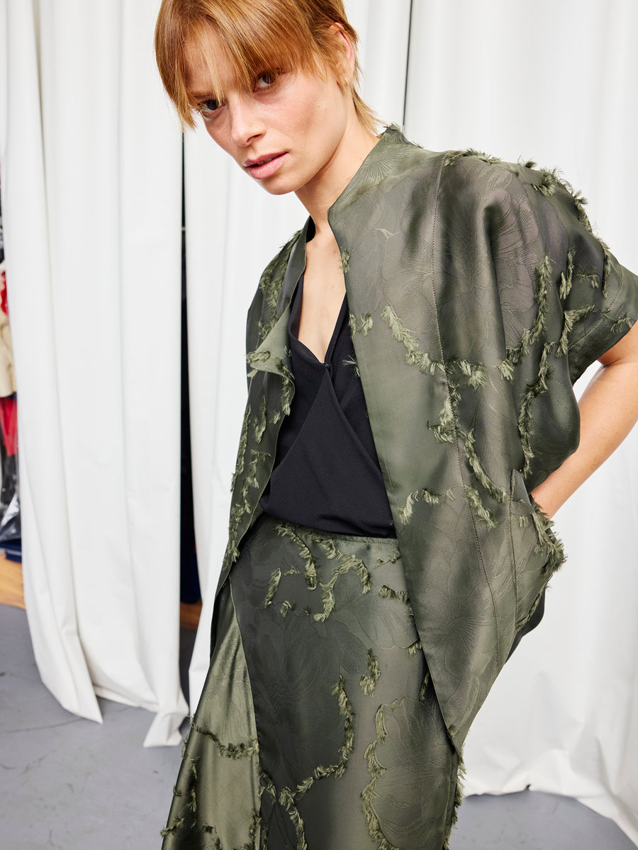 A person with short auburn hair wears an olive Ari Shrug by Zero + Maria Cornejo over a black top and matching textured two-piece, standing before white curtains and glancing slightly toward the camera.