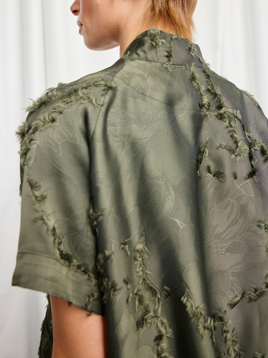A person wearing the Ari Shrug by Zero + Maria Cornejo, a short-sleeved olive green shirt with a frayed floral fil coupe texture, stands against a white curtain background, shown from the back and side.