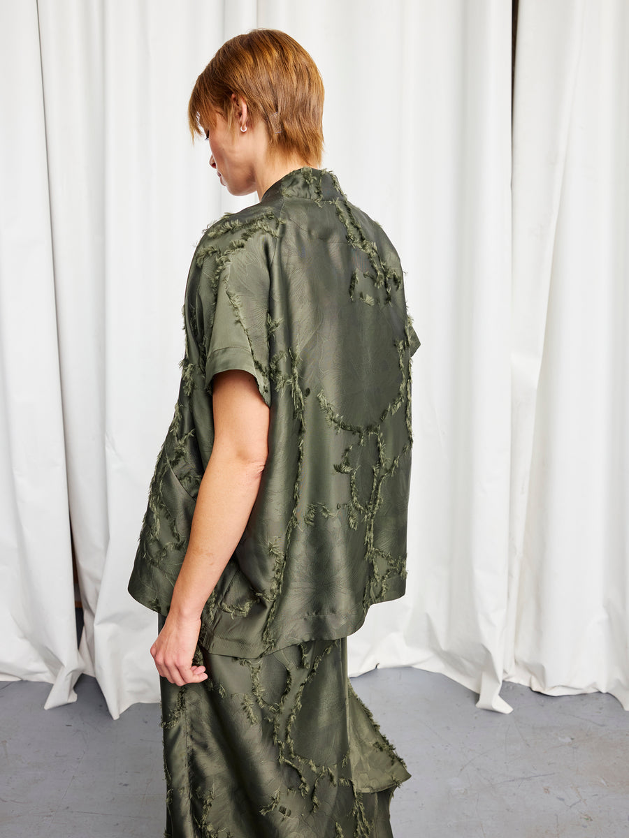 A person with short, light brown hair stands sideways in a loose olive green outfit and matching skirt, paired with the sustainable Ari Shrug by Zero + Maria Cornejo. White curtains are visible in the background.