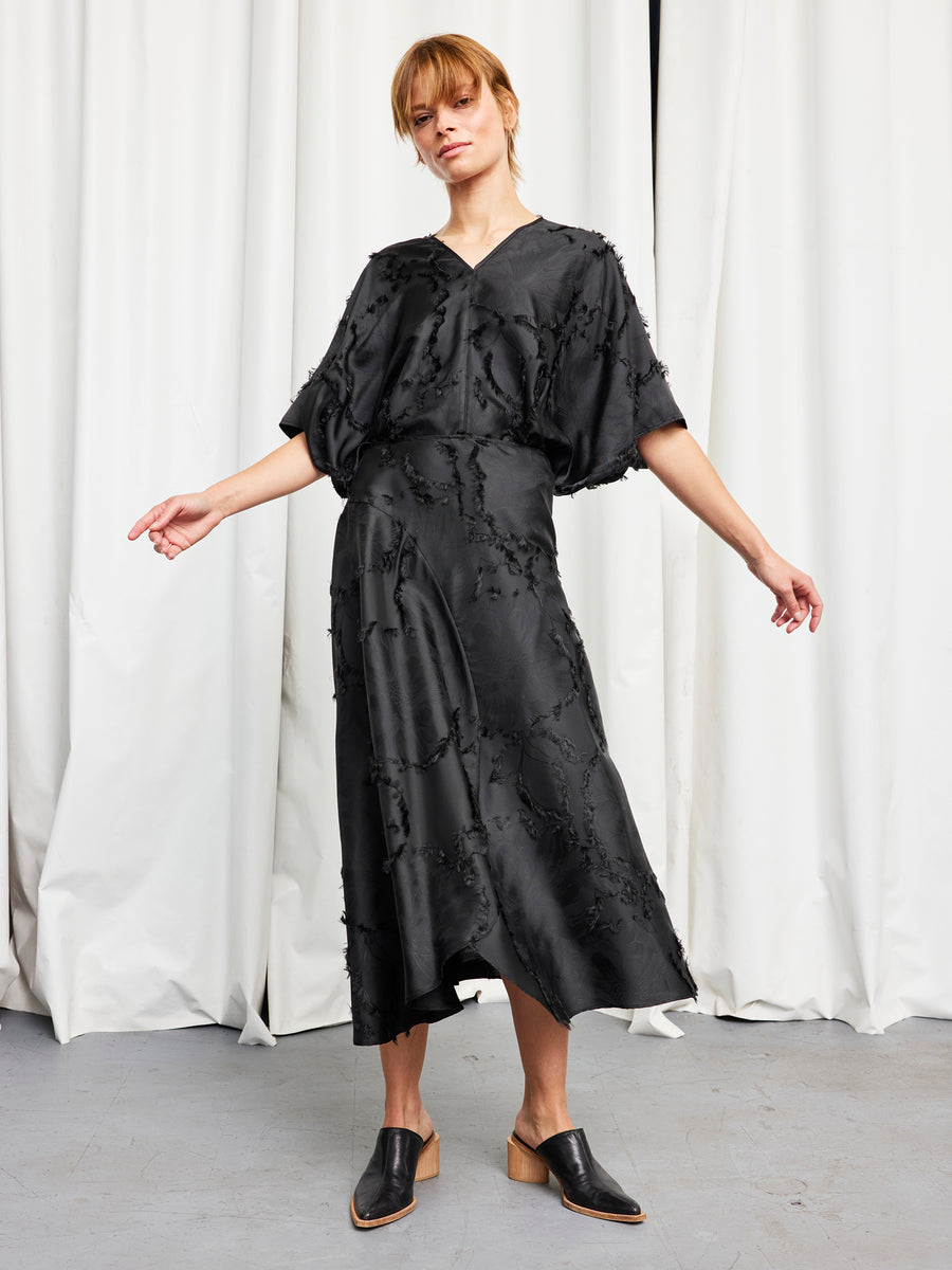 A woman stands before white curtains in a loose black midi dress with elbow-length sleeves and black mule shoes. Her look is elevated by the Zero + Maria Cornejo Circle Skirt, crafted from sustainable FSC viscose floral jacquard.
