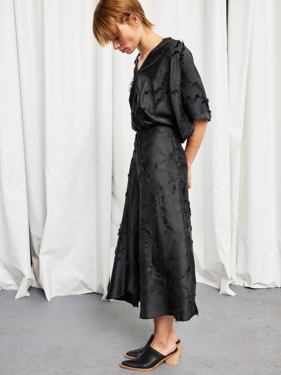 A person with short hair stands in profile, wearing a black Circle Skirt by Zero + Maria Cornejo and black mule shoes with a low wooden heel, against a white curtain backdrop.