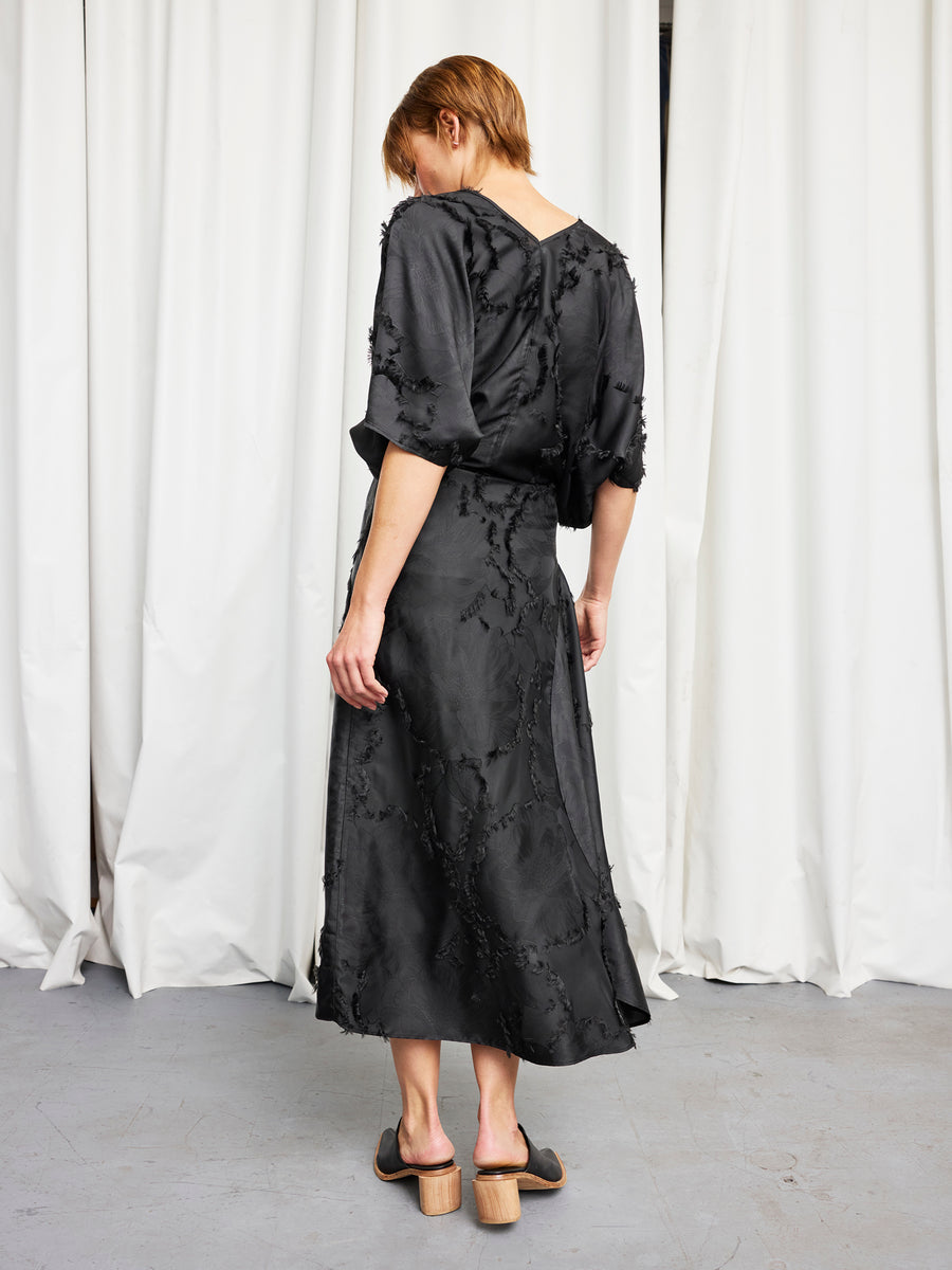 A person with short hair stands facing away, wearing the black textured Circle Skirt from Zero + Maria Cornejo with heeled sandals. Crafted from FSC viscose for sustainability, white curtains and a gray floor set the scene.