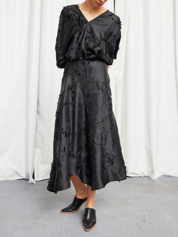 A woman stands on a gray floor before white curtains, hands behind her back and head gently lowered, wearing Zero + Maria Cornejo's black Circle Skirt with a loose black blouse and black mules.