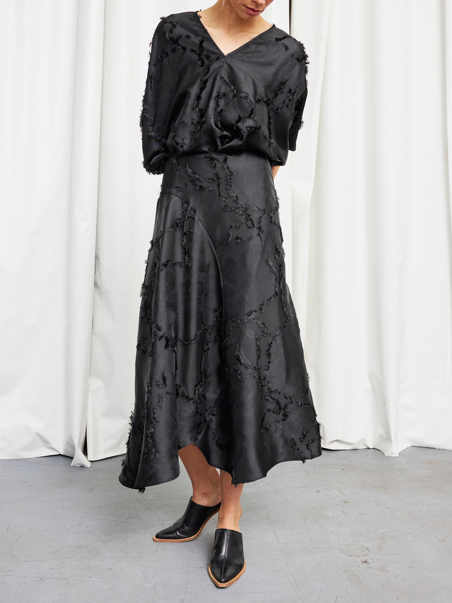 A woman stands on a gray floor before white curtains, hands behind her back and head gently lowered, wearing Zero + Maria Cornejo's black Circle Skirt with a loose black blouse and black mules.