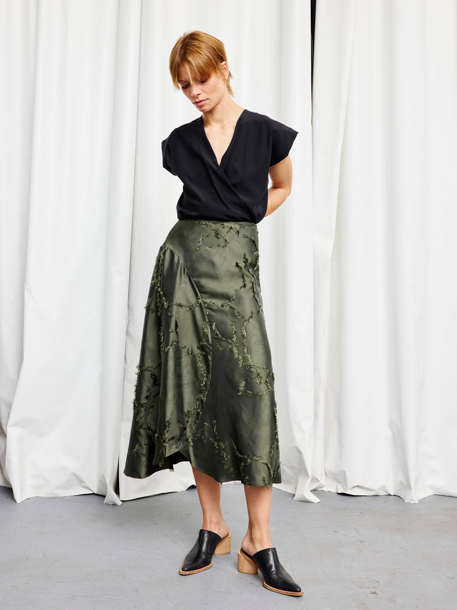A woman stands before white curtains, wearing a black short-sleeve blouse, Zero + Maria Cornejo's Circle Skirt in olive green floral jacquard, and black mule shoes. Her hands are behind her back as she gazes downward.