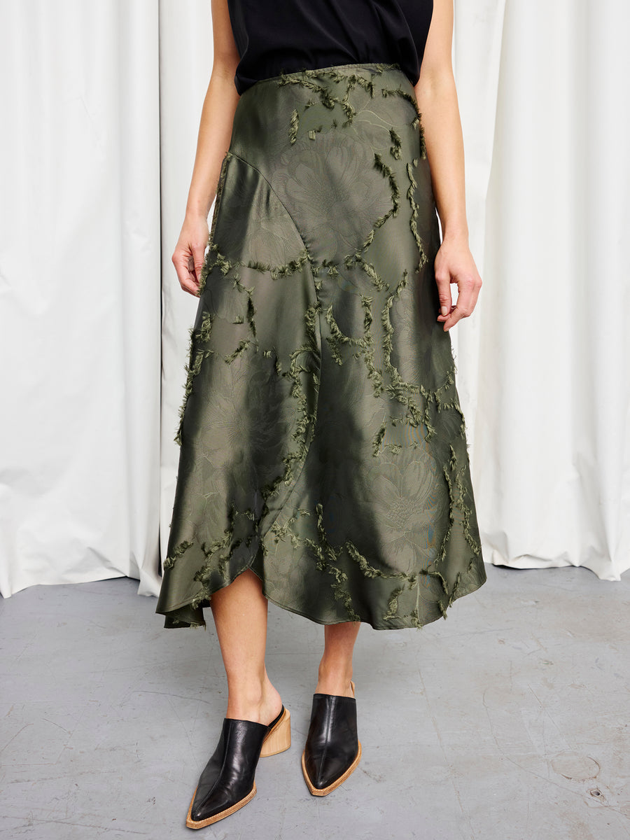 A person wearing a black sleeveless top, the olive green Circle Skirt by Zero + Maria Cornejo with a sustainable floral jacquard design, and black pointed-toe mule shoes stands in front of white curtains.