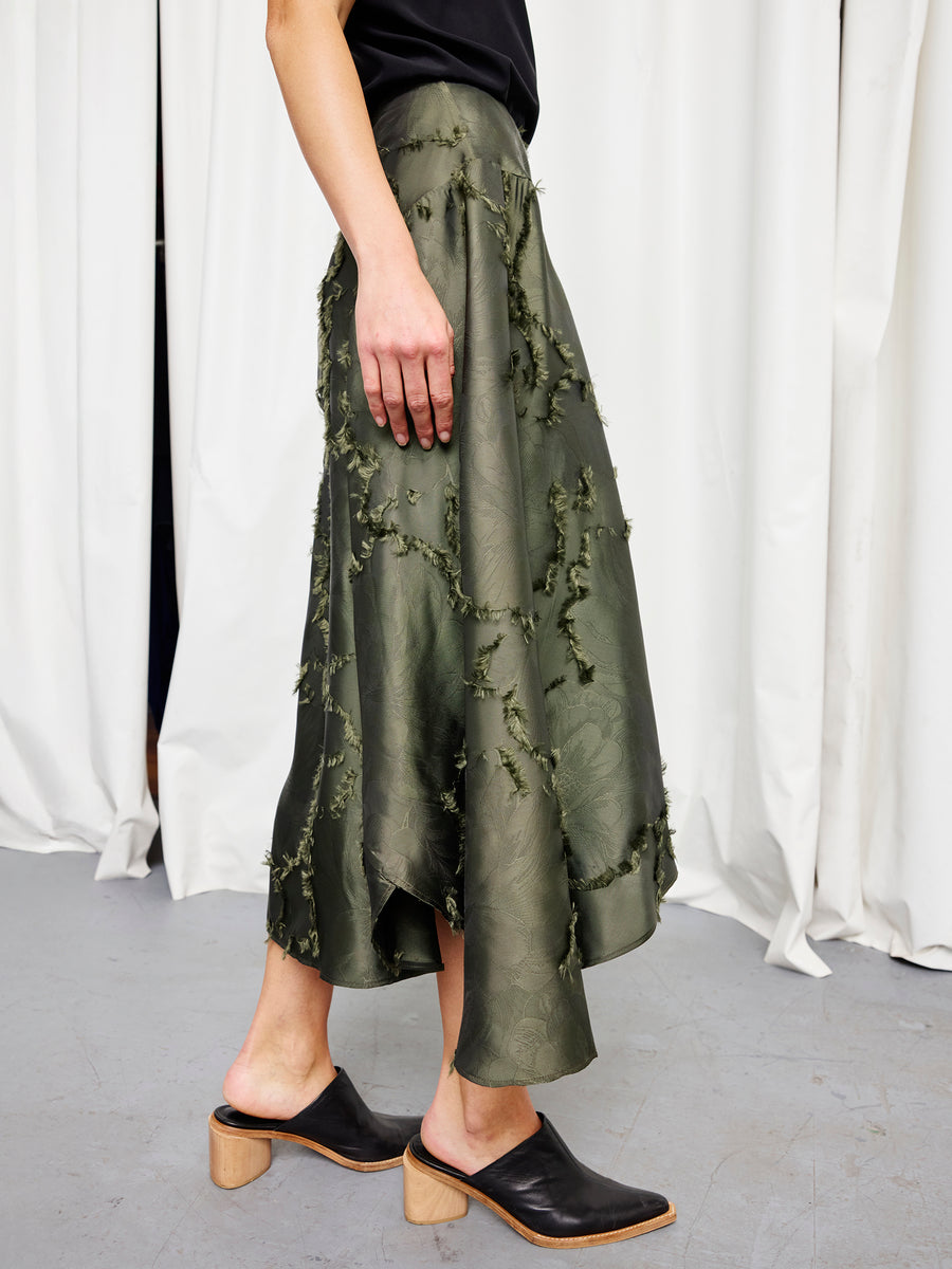 A person wears the Circle Skirt by Zero + Maria Cornejo, a shiny olive green sustainable jacquard piece with an uneven hem, styled with a black top and black wooden-heeled mules, on a gray floor before white curtains.