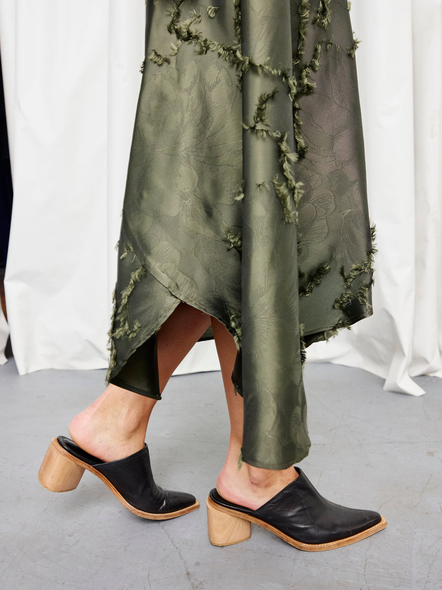 A person wears the Zero + Maria Cornejo Circle Skirt in olive green, paired with black mule heels featuring wooden block heels, standing on a gray floor before white curtains.
