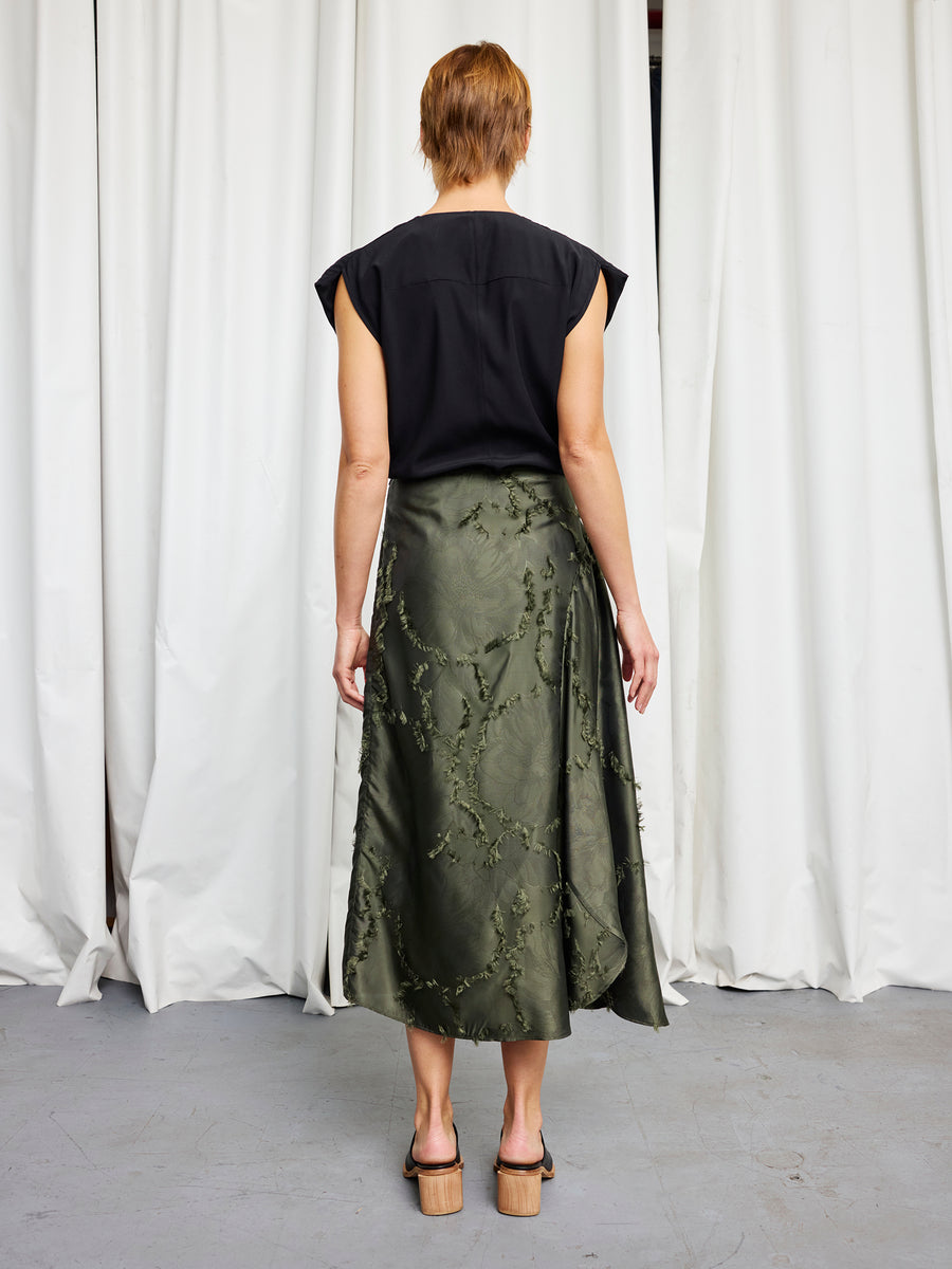A person stands with their back to the camera, wearing a black sleeveless top and the Zero + Maria Cornejo Circle Skirt in a sustainable floral jacquard, paired with heeled sandals. White curtains hang in the background.