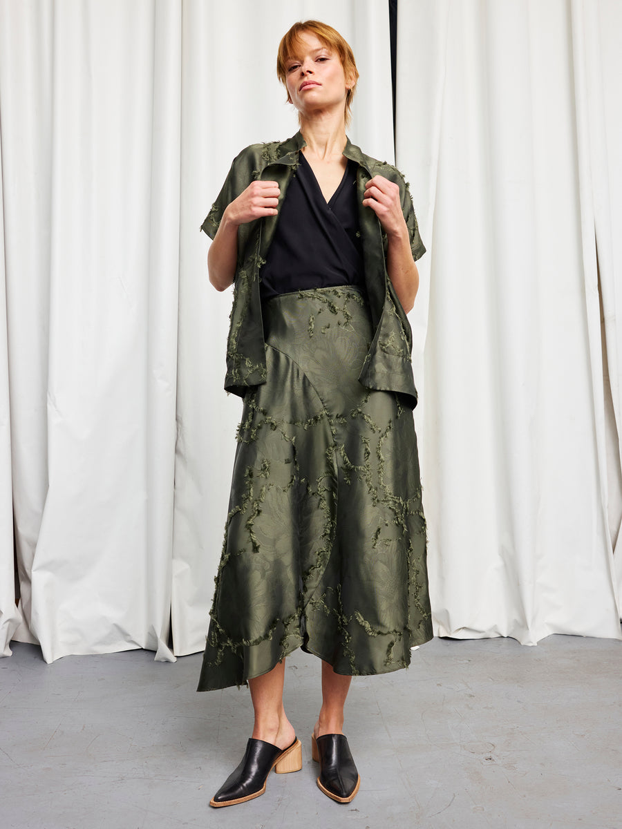 A woman stands in front of white curtains wearing the olive Circle Skirt by Zero + Maria Cornejo with a matching jacket over a black top, paired with black mules and light wooden heels.