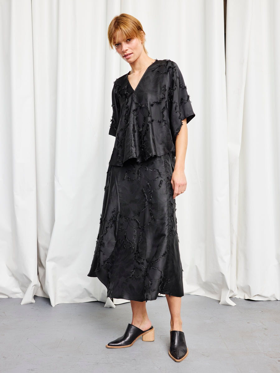 A person with short light hair stands before white curtains, wearing the Zero + Maria Cornejo Circle Top in black and a matching black midi skirt. They complete the look with black mule shoes featuring a wooden heel.