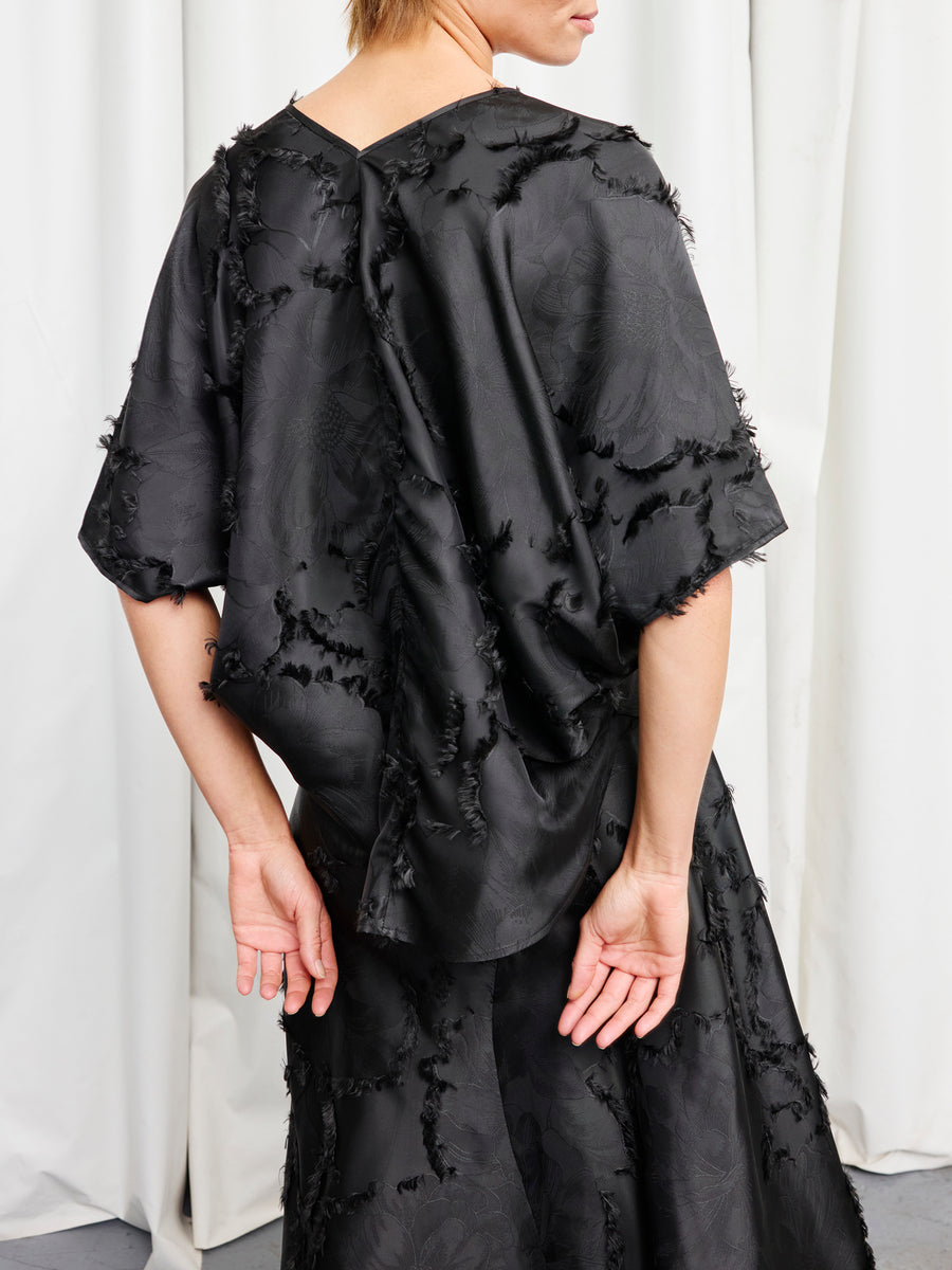 A person stands with their back to the camera, wearing the Circle Top by Zero + Maria Cornejo—a loose, sustainable floral jacquard top paired with a matching skirt. One arm is bent behind them; white curtains hang in the background.