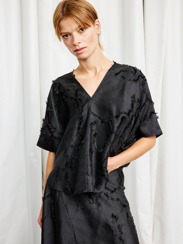 A woman with light brown hair wears the Circle Top by Zero + Maria Cornejo—a loose, short-sleeved sustainable jacquard top with textured, frayed details and a subtle v-neck—paired with a matching skirt in front of white curtains.