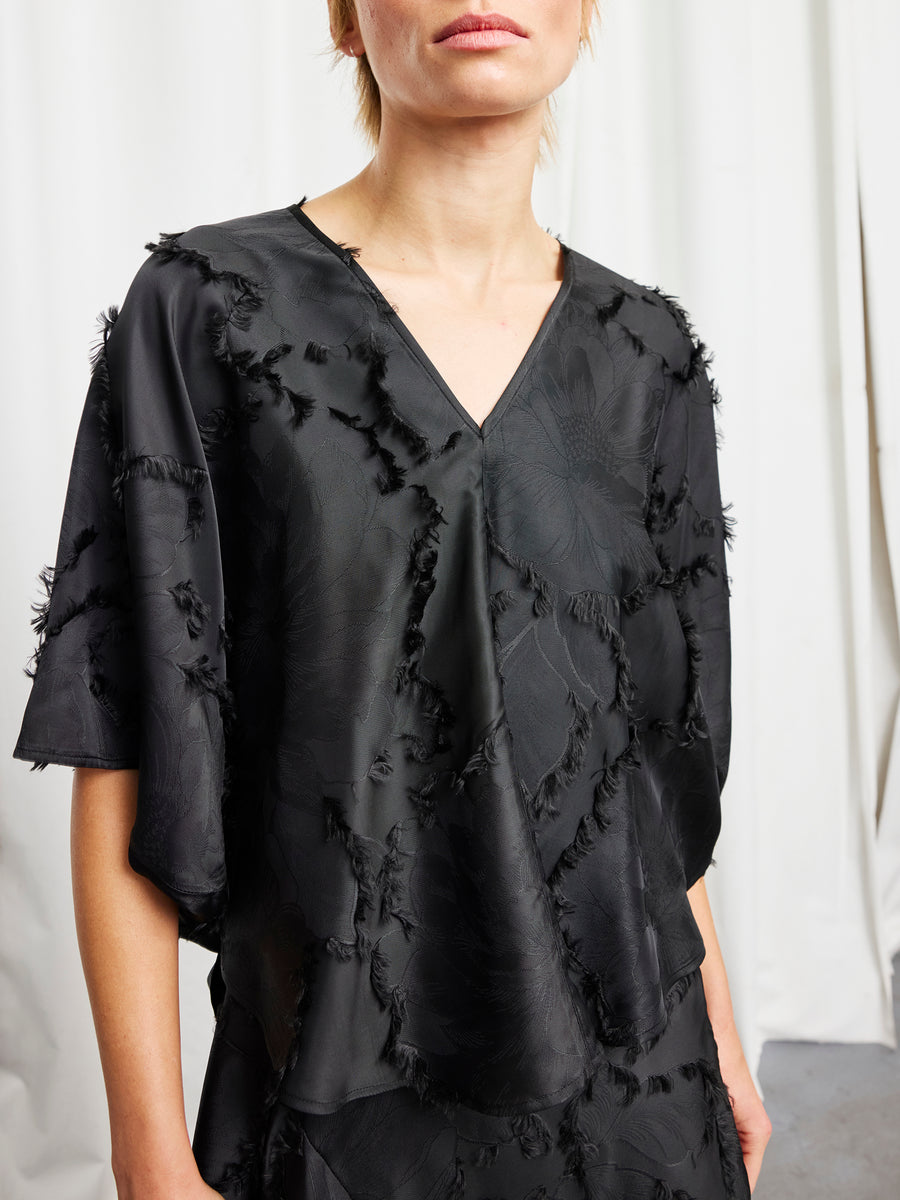 A person with short blond hair, face partially visible, stands before white curtains wearing the Circle Top by Zero + Maria Cornejo—a loose-fit sustainable jacquard piece with a V-neck and frayed floral patterns.
