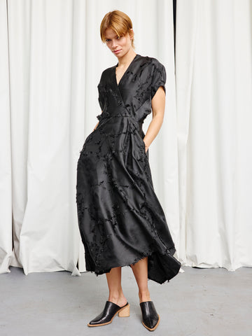 A person in the black Long Aki Wave Dress by Zero + Maria Cornejo and black mule heels stands confidently before white curtains, hands in pockets, looking at the camera.