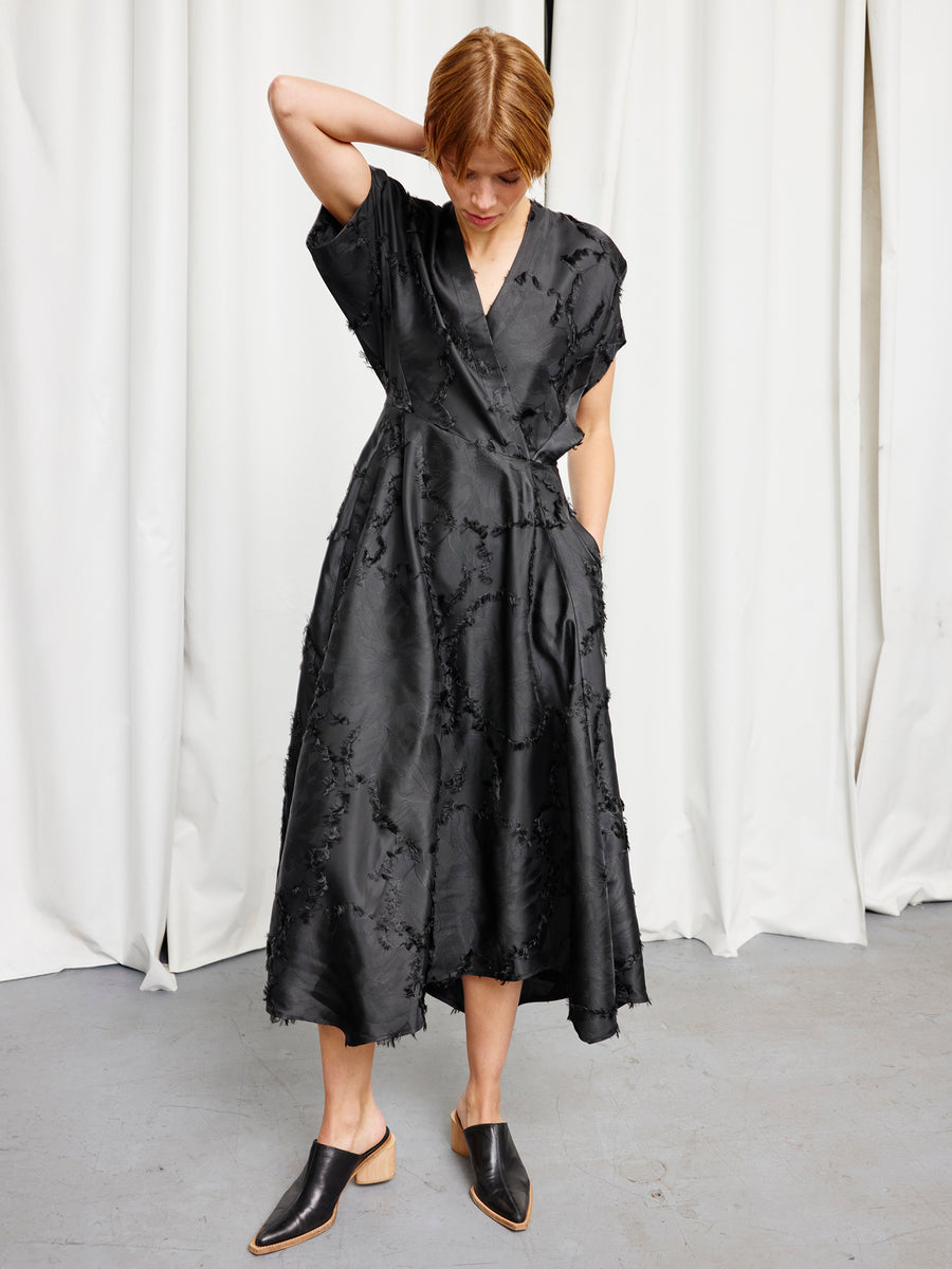 A woman stands indoors by white curtains, wearing the black Long Aki Wave Dress by Zero + Maria Cornejo, featuring a textured pattern and loose fit. She looks down with one hand on her head, paired with black mule shoes.