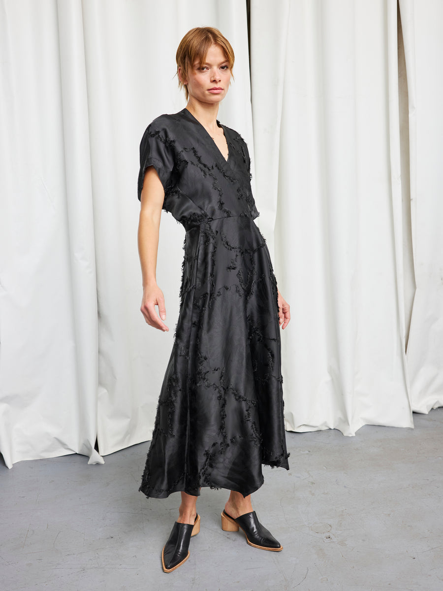 A woman stands indoors before white curtains, wearing the Zero + Maria Cornejo Long Aki Wave Dress—black fil coupé jacquard with short sleeves and v-neck—paired with black mule shoes featuring wooden heels.
