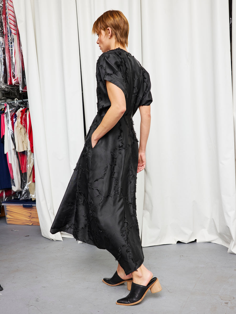 A person with short hair stands sideways, wearing the Zero + Maria Cornejo Long Aki Wave Dress with short sleeves and black heeled mules. One hand is in their pocket. Behind them are hanging clothes and white curtains.