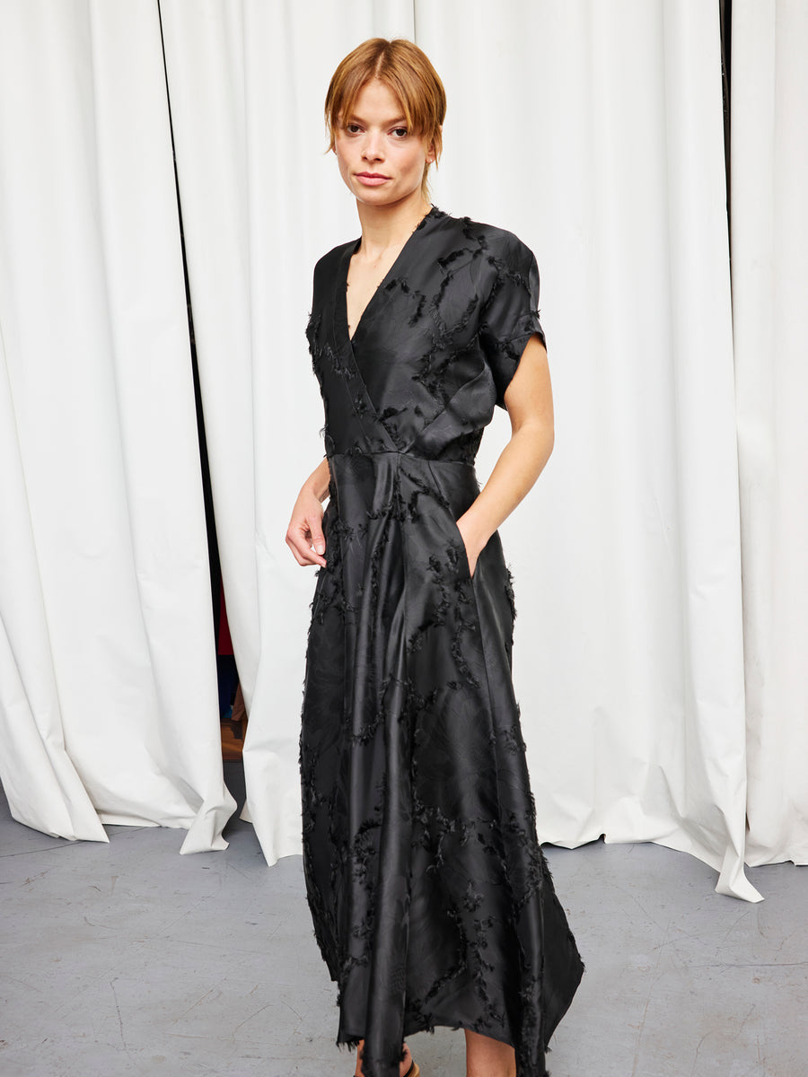 A person stands indoors by white curtains, confidently wearing the Zero + Maria Cornejo Long Aki Wave Dress—a black, short-sleeved fil coupé jacquard dress with pockets—while looking at the camera with a relaxed posture.