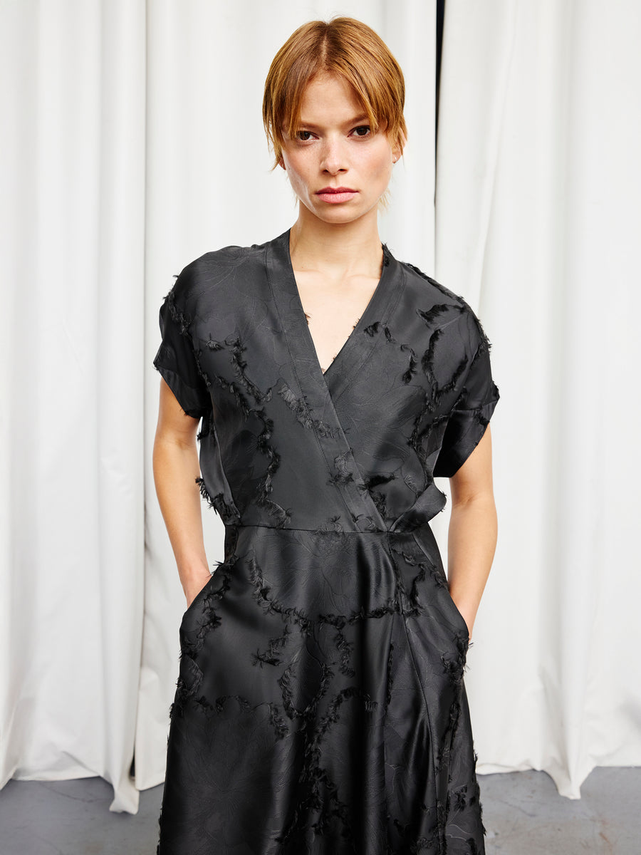 A person with short, light brown hair models the Zero + Maria Cornejo Long Aki Wave Dress—a textured black, full-length wrap dress with short sleeves—standing in front of white curtains, hands in their pockets, looking at the camera.