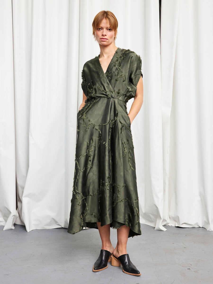 A person with short, light brown hair stands before white curtains wearing the Zero + Maria Cornejo Long Aki Wave Dress—a textured, dark green wrap dress with short sleeves and a v-neck—paired with black mule shoes and hands in the pockets.