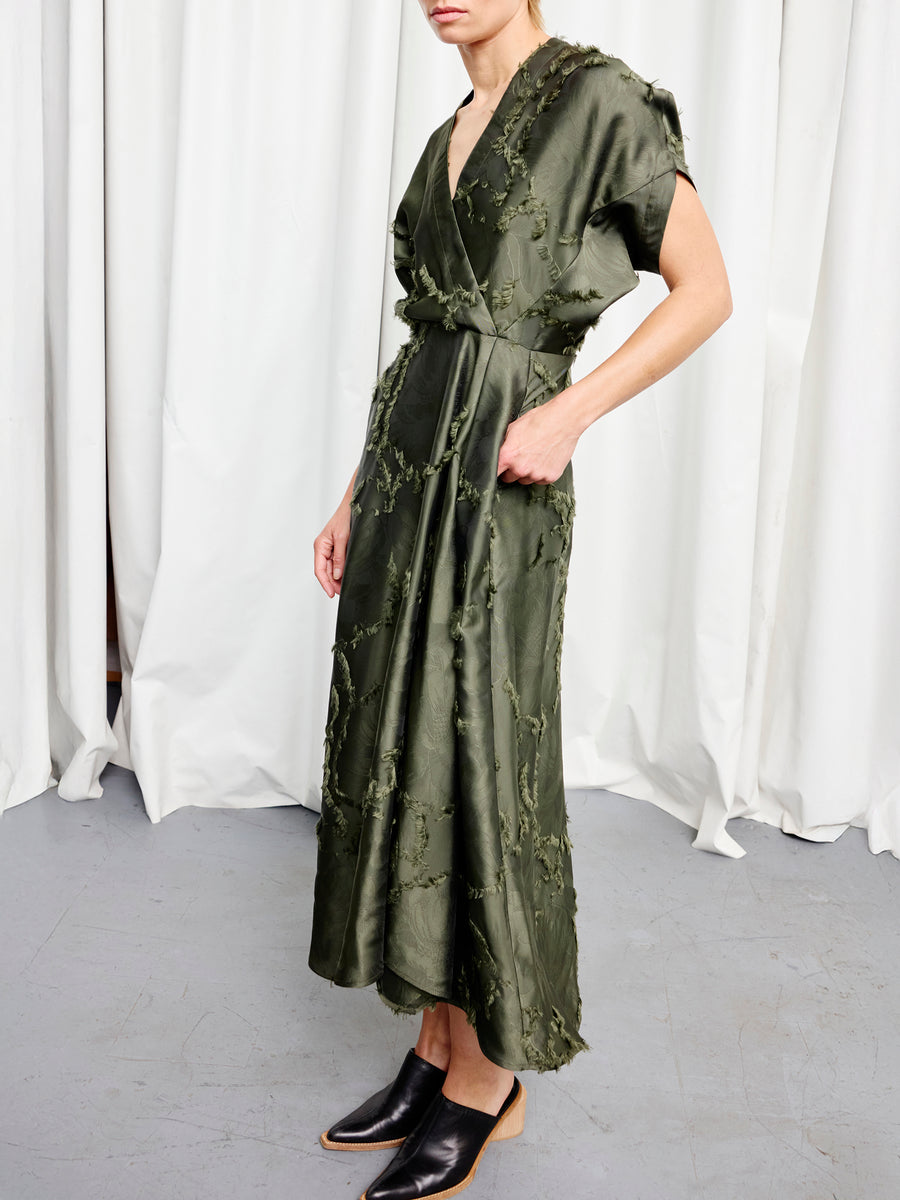 A woman stands against white curtains in the Zero + Maria Cornejo Long Aki Wave Dress—an olive green, fil coupé jacquard wrap style with textured floral details—her face partially out of frame, wearing black backless mules.