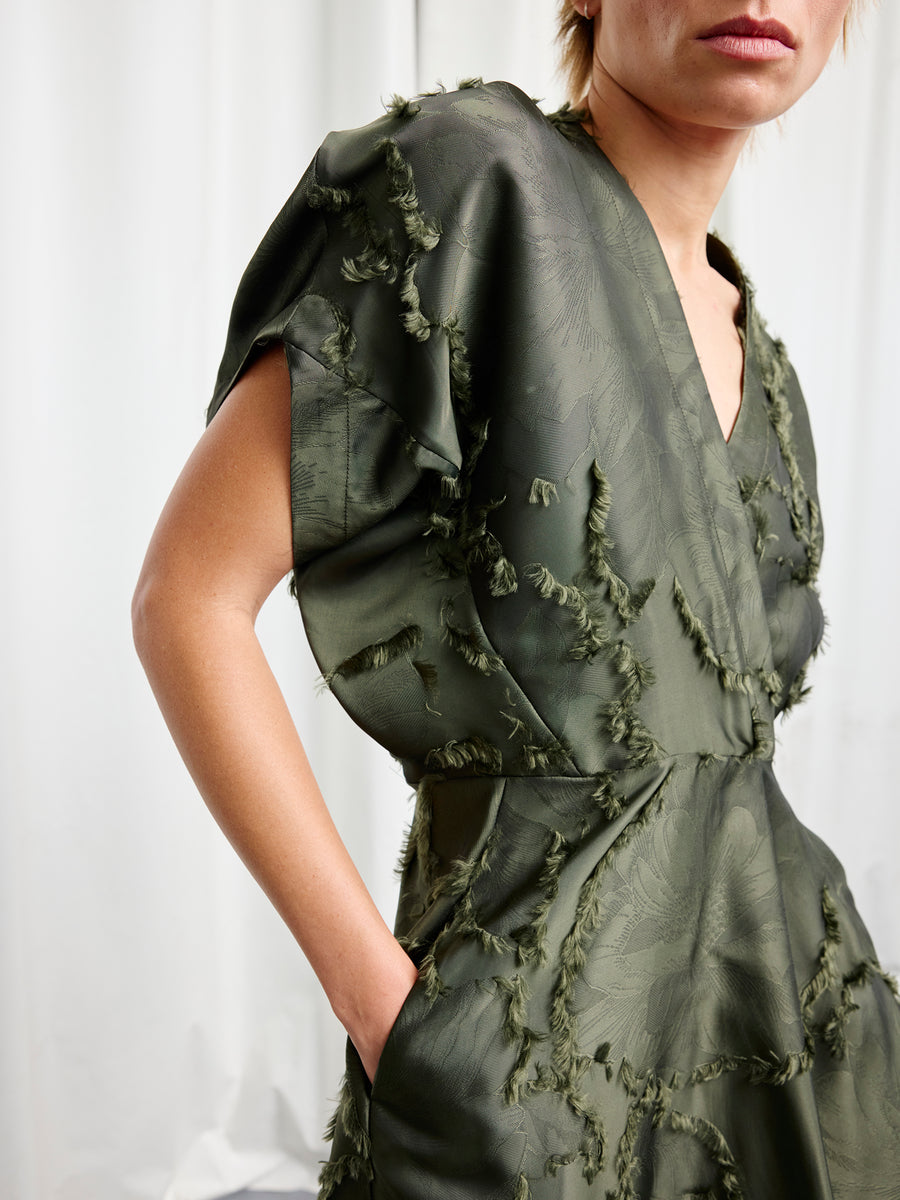 A person models the Zero + Maria Cornejo Long Aki Wave Dress—a green, textured fil coupé jacquard piece with frayed edges and a V-neck—standing with one hand in a pocket against a white background. The image shows their upper body and part of their face.