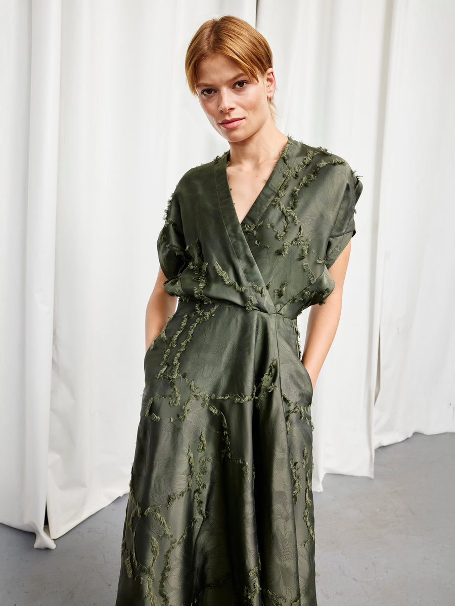 A woman stands indoors wearing the Zero + Maria Cornejo Long Aki Wave Dress in olive, a textured, full-length wrap dress with short sleeves and pockets. White curtains are in the background.