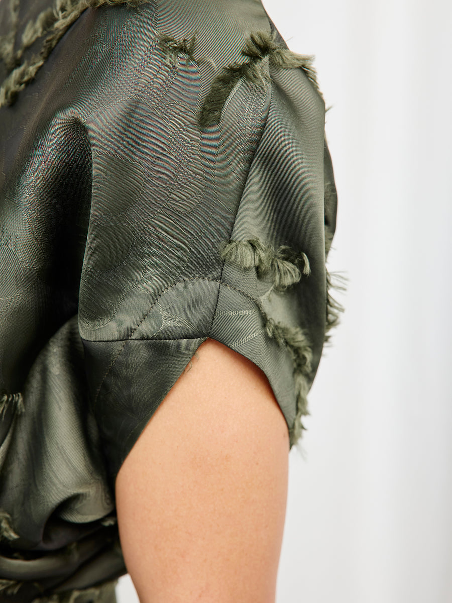 A close-up of a person's upper arm in Zero + Maria Cornejo's Long Aki Wave Dress, featuring dark green satin-like fabric with frayed seams and a subtle floral pattern. The background is plain and light-colored.
