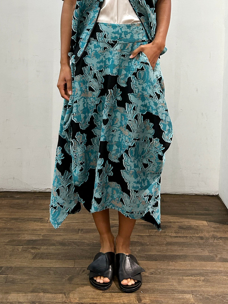 A person stands on a wood floor in a patterned turquoise and black outfit. The asymmetrical Nio Skirt by Zero + Maria Cornejo, made from sustainable jacquard, pairs with the matching vest. One hand rests in a pocket above sleek black slide sandals, exuding effortless style.