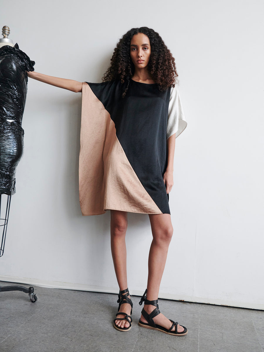 A woman with curly hair models the Dia Dress by Zero + Maria Cornejo, a linen-blend garment with a color-block design in black, beige, and white sections. She poses with one arm resting on a dress form, wearing strappy sandals against a plain white background.