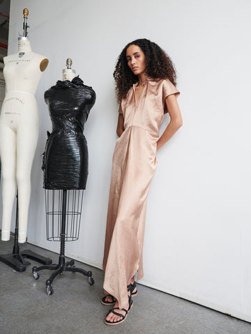 A woman with curly hair stands next to two dress forms in what resembles a design studio. She is wearing black sandals and the Long Ruched Stella Dress by Zero + Maria Cornejo, which is a long, silky, light-colored v-neck dress. One dress form holds a black textured piece, while the other displays a plain design.