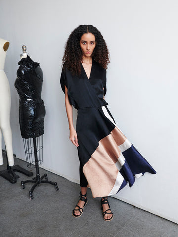 A person with curly hair stands in a chic ensemble featuring the Patchwork Aki Wave Dress by Zero + Maria Cornejo, showcasing an asymmetrical design with flowing panels in black, beige, white, and navy. They are positioned in a studio beside two dress forms, one draped in black plastic.