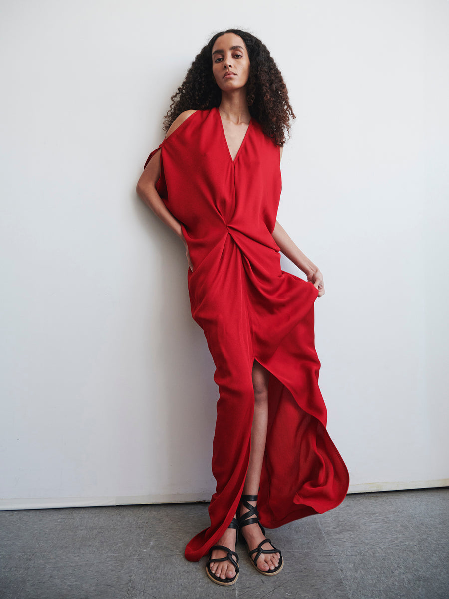 A person with curly hair poses against a white wall, wearing the Long Miu Dress by Zero + Maria Cornejo. The sustainably-sourced dress, made of woven viscose, is vibrant red and drapes elegantly down to the floor, featuring a deep V-neck and front slit. They stand barefoot in black sandals.