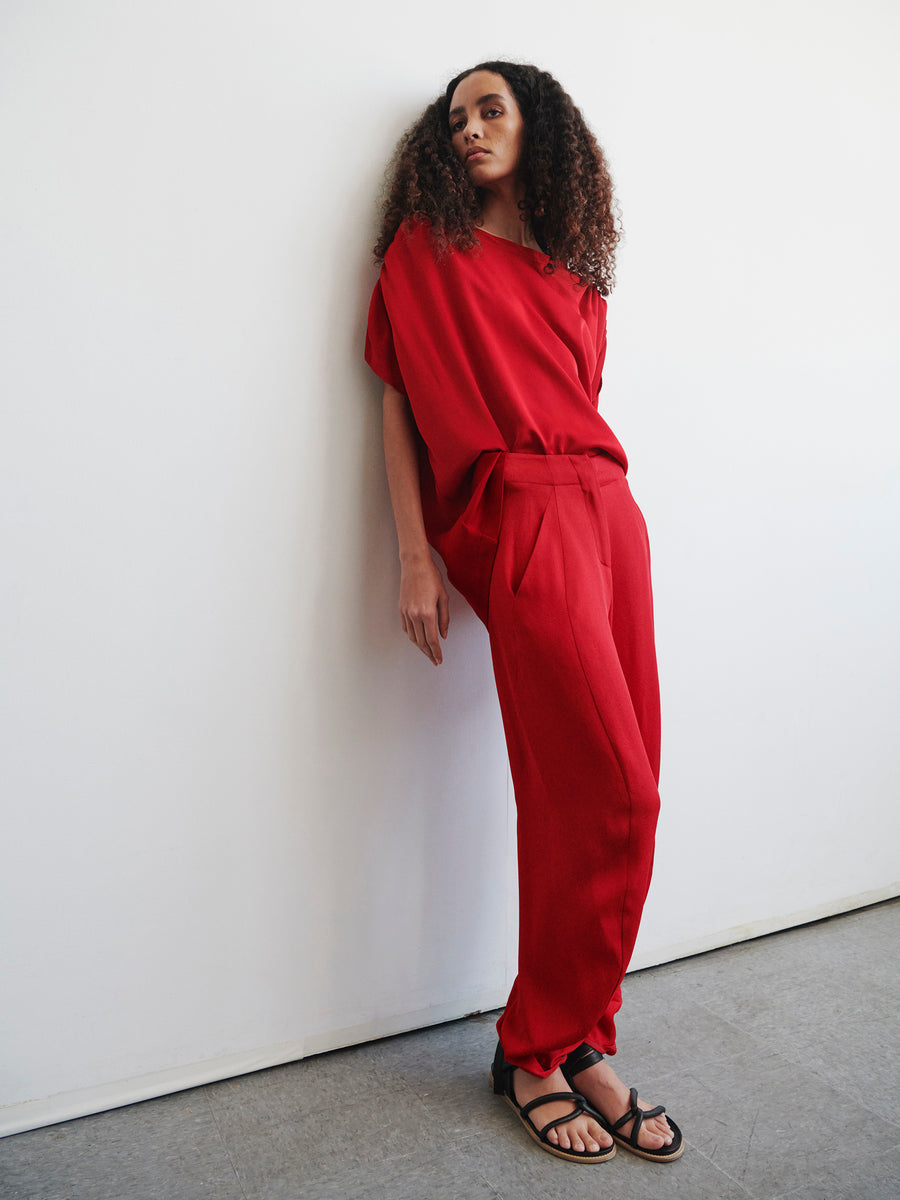 A person with curly hair, wearing the Takeo Pant in eco drape red from Zero + Maria Cornejo paired with black sandals, leans against a white wall on a gray floor while gazing slightly upward. This outfit showcases sustainably-sourced materials for those who value stylish yet responsible fashion.