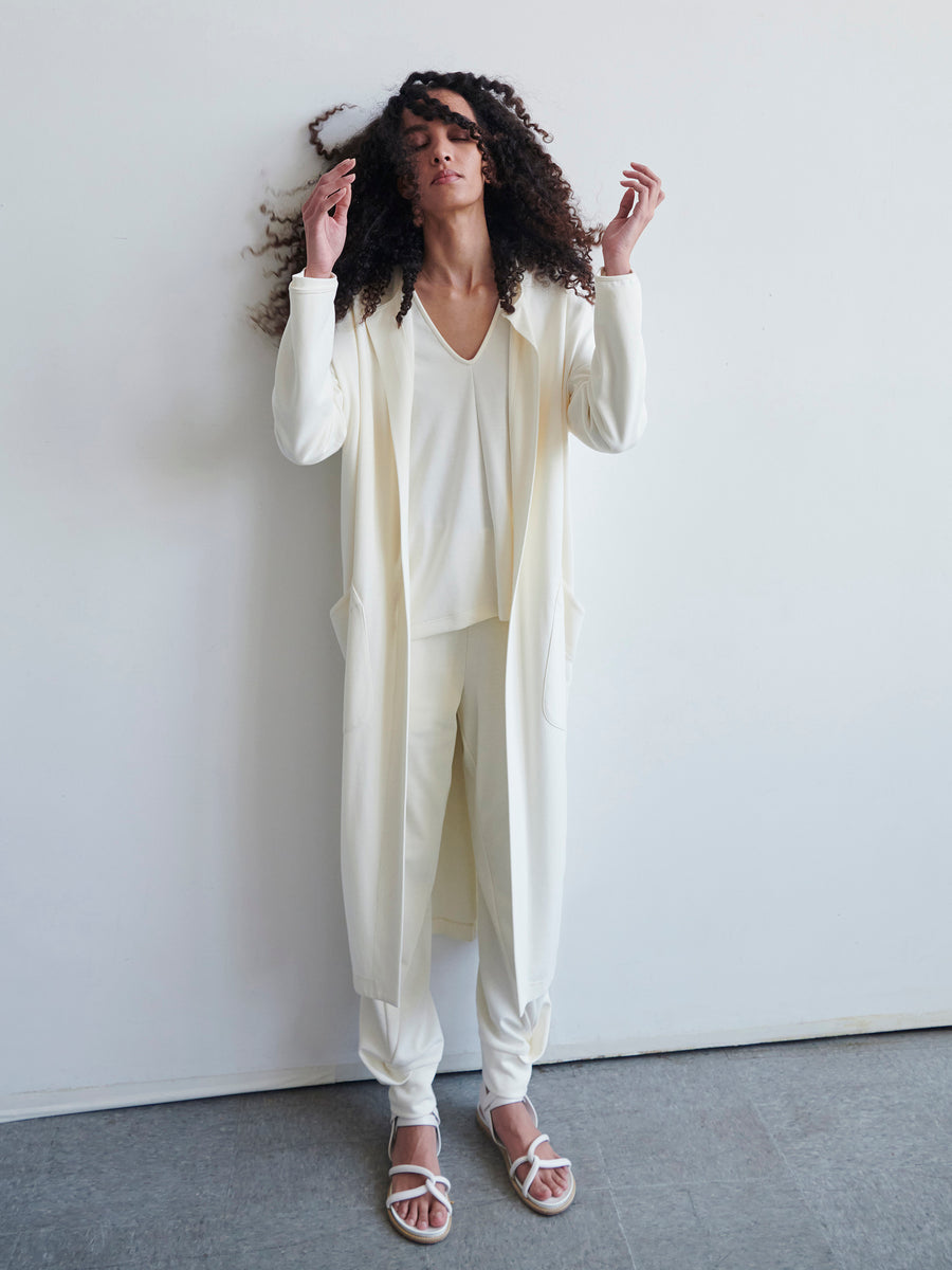 A person with long curly hair exudes elegance in a white ensemble made from cotton-modal jersey, featuring the Jersey Koya Coat by Zero + Maria Cornejo. The outfit, showcasing a cocoon silhouette with a cardigan, top, and pants, is effortlessly complemented by white sandals as they pose against a plain white wall with hands gently raised.