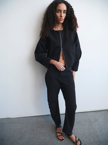 A person with long curly hair stands against a white wall, wearing the T Bomber by Zero + Maria Cornejo paired with black pants and black sandals. They exude a relaxed posture, one foot slightly forward and their hands casually tucked into pockets.