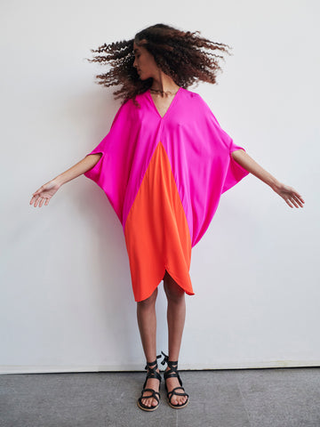 A person stands against a white wall wearing the vibrant Koya Dress by Zero + Maria Cornejo, featuring pink and orange silk charmeuse panels. Their arms are outstretched, and their curly hair is caught in motion. They are wearing black sandals.